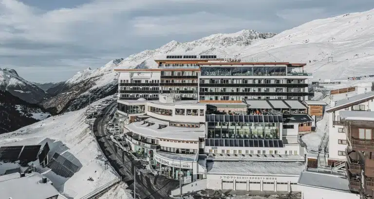 Modernes mehrstöckiges Hotel in verschneiten Bergen mit kurvenreicher Straße und bewölktem Himmel.