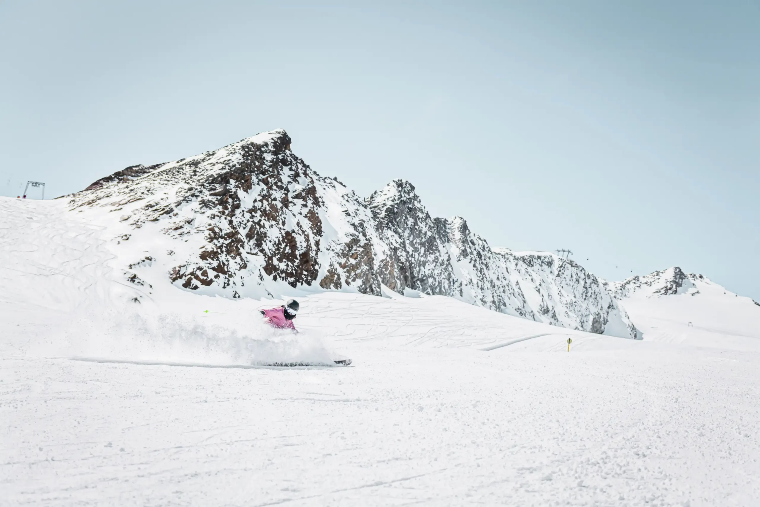 Skifahrer in rosafarbener Jacke gleitet auf einem verschneiten Berg unter klarem Himmel bergab.