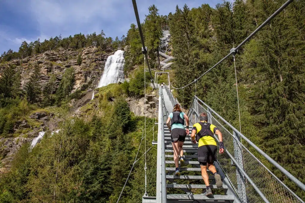 Zwei Personen erklimmen auf einem Trailrunning im Ötztal eine Außentreppe, die an einem Wasserfall und felsigen Hügeln vorbeiführt.