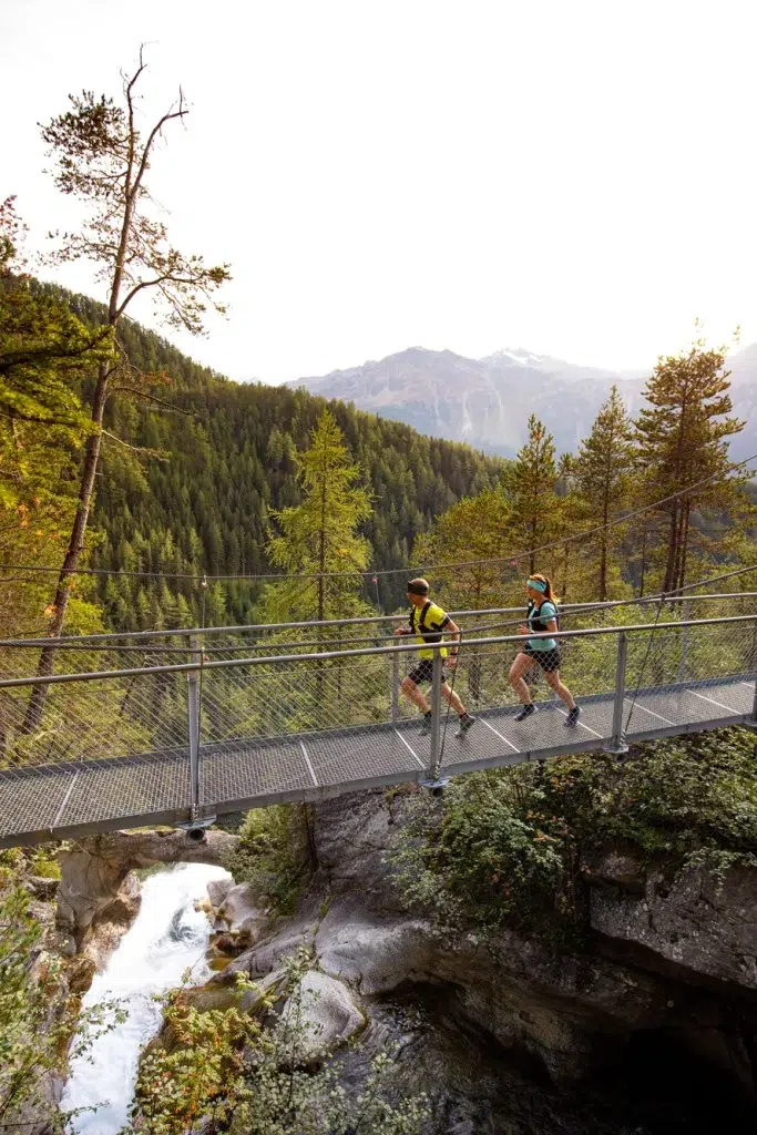 Zwei Personen genießen Trailrunning im Ötztal über eine Brücke oberhalb einer bewaldeten Schlucht und Berge.
