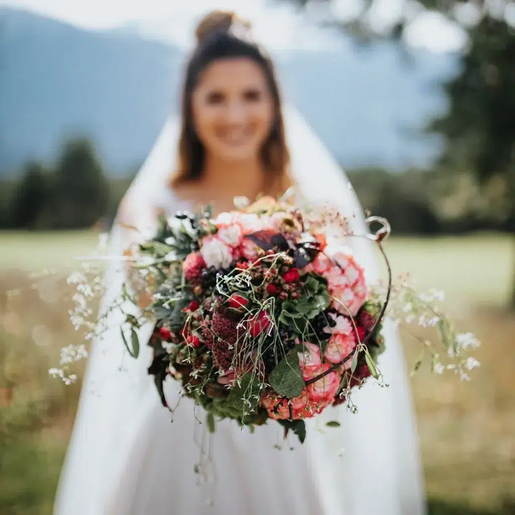 Die Braut im Freien auf der Heiraten Stöttlalm mit einem farbenfrohen Strauß aus grünen und rosa Blumen.