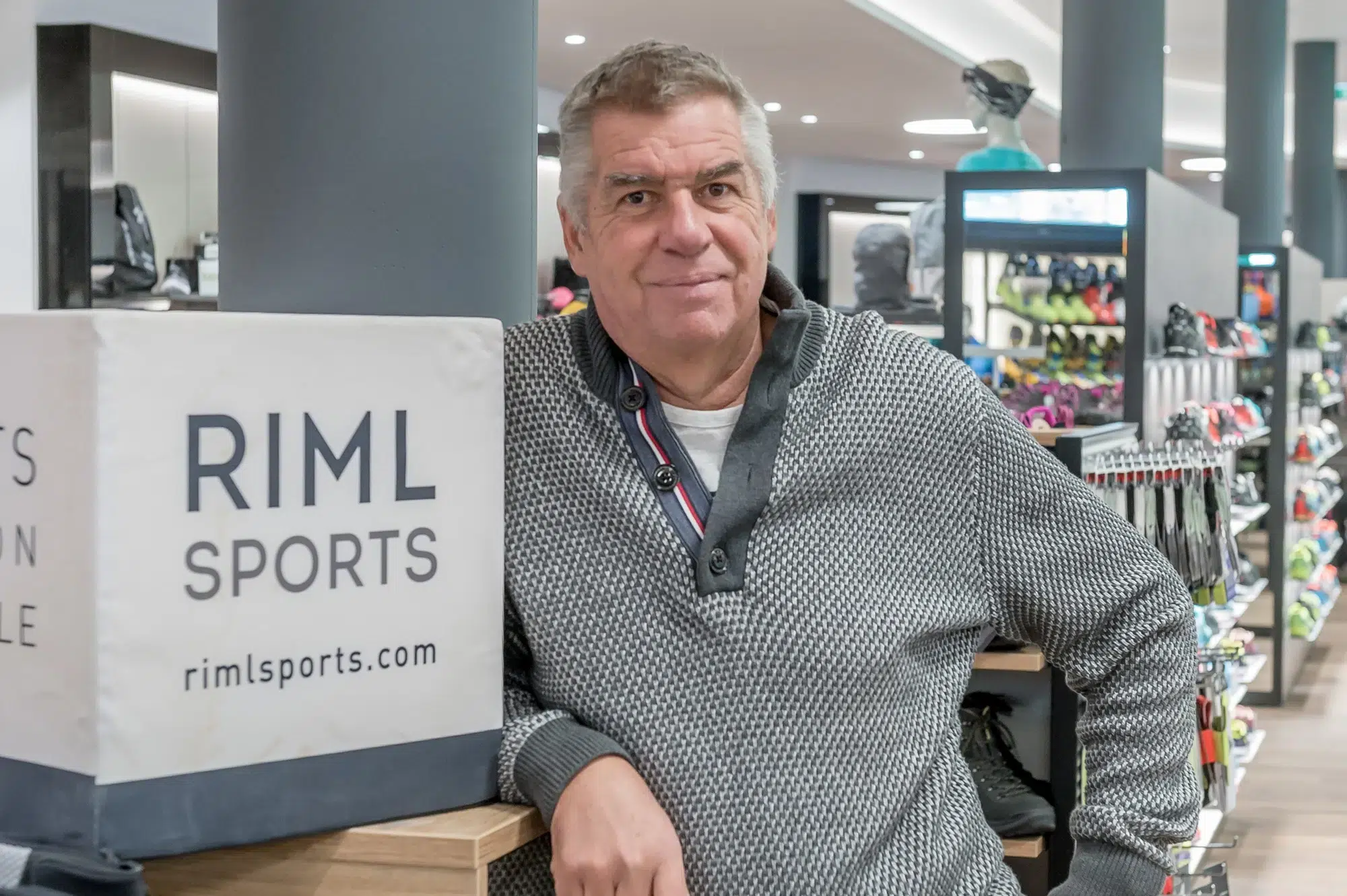 Ein Mann steht in einem Sportgeschäft neben einem Schild mit der Aufschrift RIML SPORTS rimlsports.com, Piccard No5.