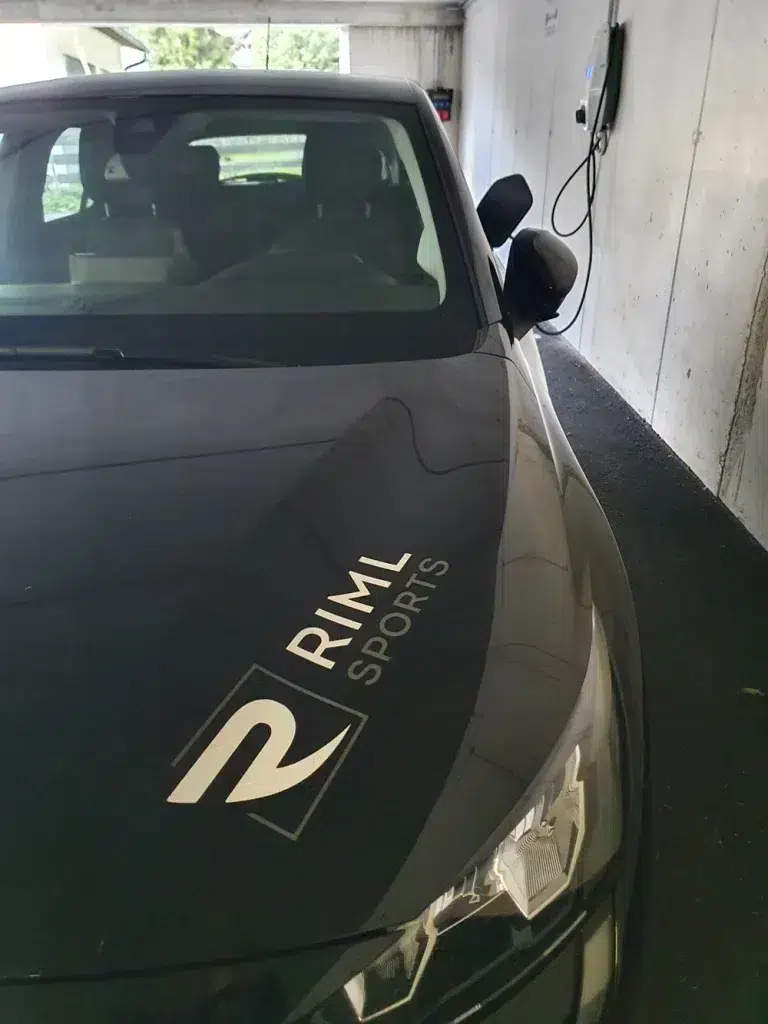 Ein schwarzes Auto mit RML SPORTS-Logo, das für Nachhaltigkeit im Sporthandel wirbt, an einer Ladestation.