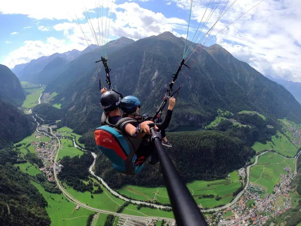 Zwei Personen genießen einen Fly Airea Tandem-Gleitschirmflug über einem üppig grünen Tal und den Bergen.
