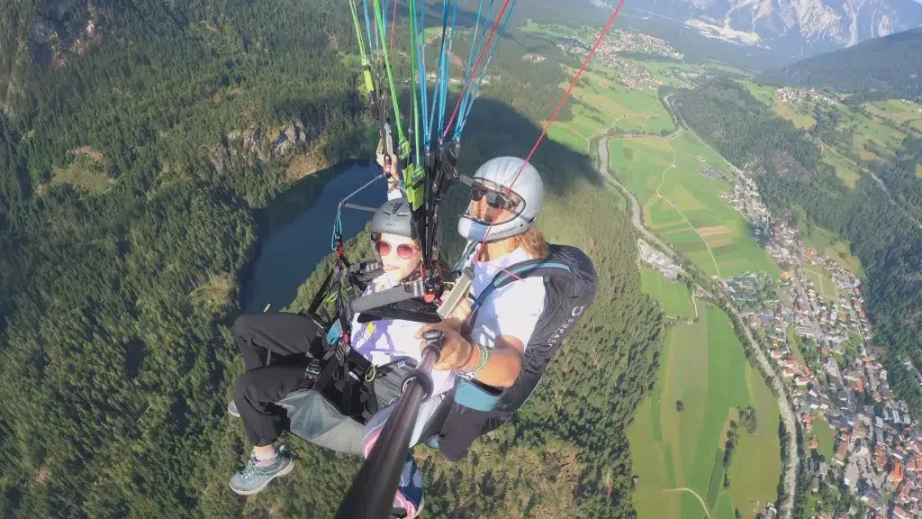 Zwei Personen fliegen beim Airea Tandem-Paragliding über eine grüne Landschaft und machen ein gemeinsames Selfie.