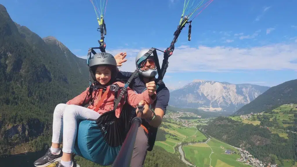 Zwei Personen genießen einen Fly Airea Tandem-Gleitschirmflug über einem grünen Tal und lächeln in die Kamera.