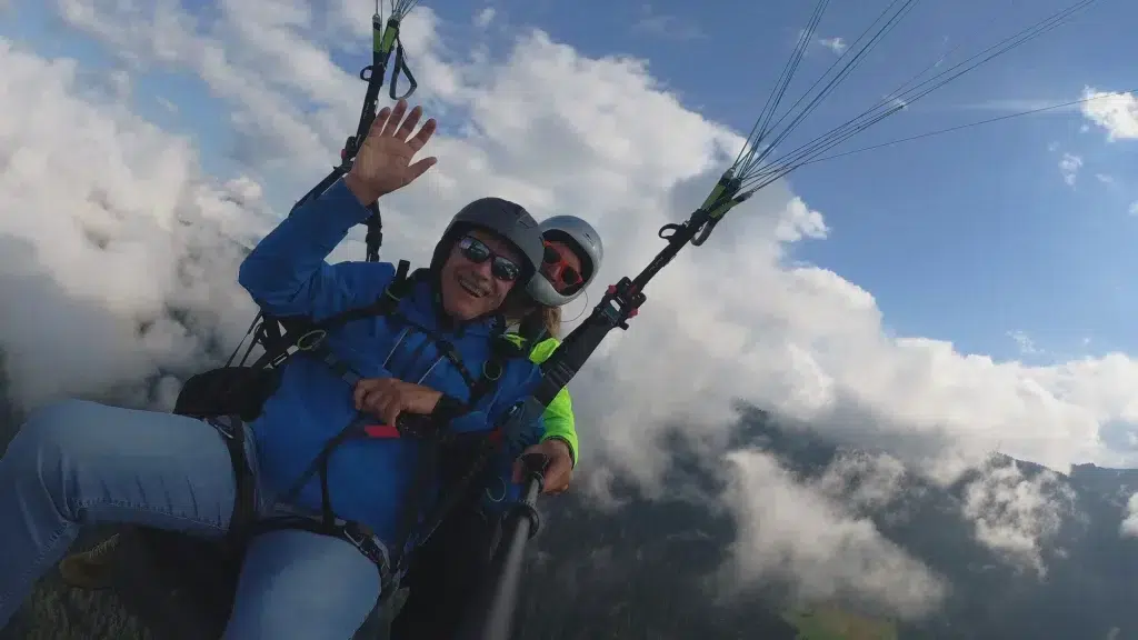 Zwei Personen genießen ein Fly Airea Tandem-Paragliding-Abenteuer über den Wolken, einer winkt und lächelt.