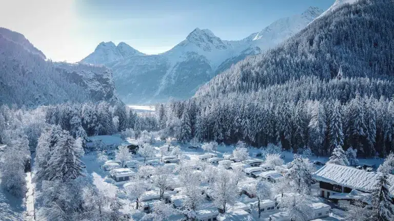 Schneebedeckte Hütten zwischen Bäumen auf dem Camping Ötztal, Berge, die sich in den klaren Himmel erheben.