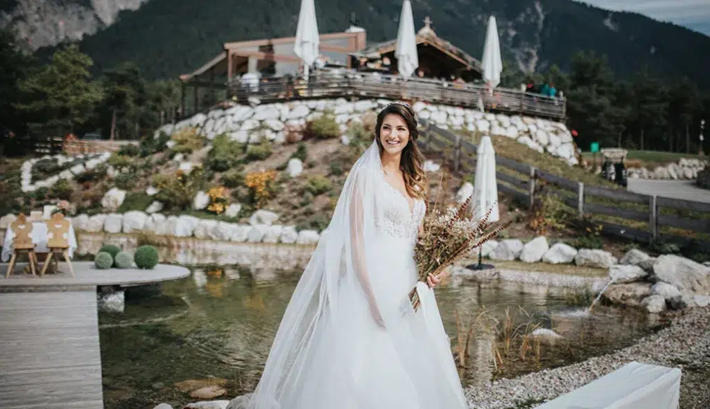 Braut in weißem Kleid mit Blumenstrauß, lächelnd im Freien auf der Heiraten Stöttlalm mit Bergblick.