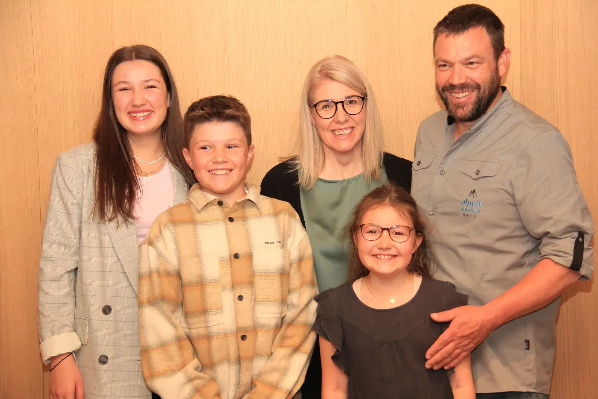 Eine lächelnde fünfköpfige Familie posiert gemeinsam vor dem hölzernen Hintergrund des Hotels Alpenaussicht.