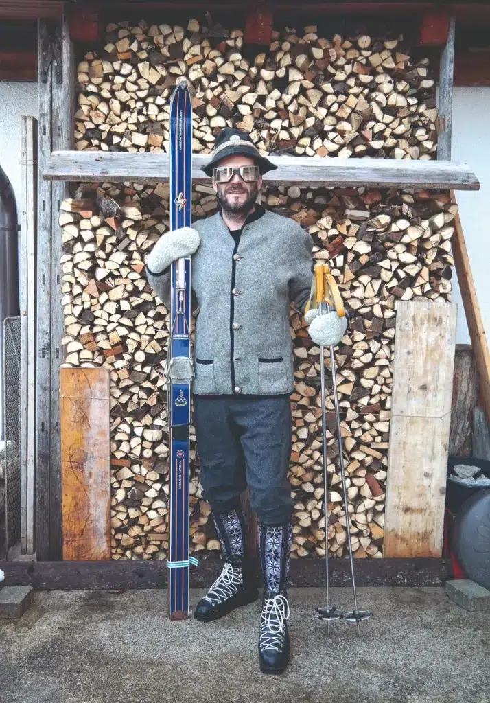 Skiinfluencer in traditioneller Kleidung posiert mit alten Skiern vor einem Stapel Brennholz.
