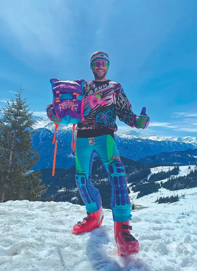 Skiinfluencer in bunter Ausrüstung steht auf einem verschneiten Berg mit blauem Himmel und Gipfeln im Hintergrund.