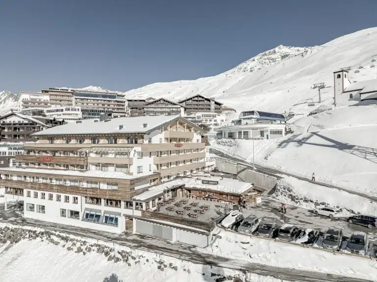 Verschneites Skigebiet mit Sporthotel Olymp, Chalets, geparkten Autos und Bergen unter klarem Himmel.