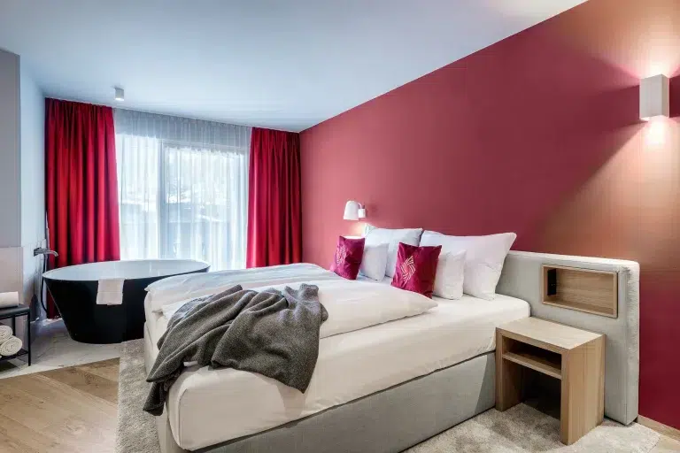 Modernes Hotelzimmer mit einer roten Akzentwand, einem großen Bett und der schwarzen Badewanne von The Secret am Fenster.