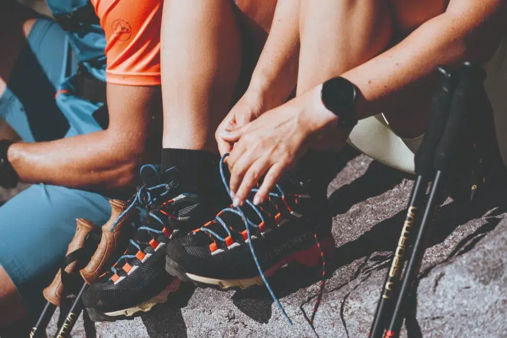 Person, die auf einem Felsen sitzend ihre Wanderschuhe bindet, während in der Nähe Trekkingstöcke und Beine zu sehen sind.