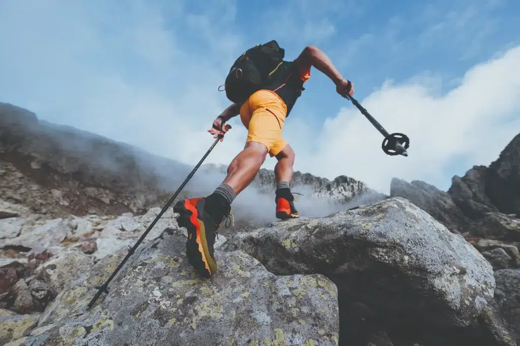 Ein Wanderer in orangefarbenen Shorts erklimmt mit Trekkingstöcken felsiges Gelände unter blauem Himmel.