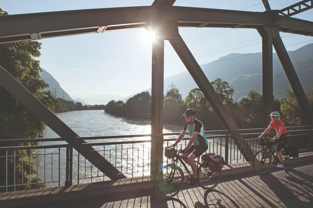 Zwei Personen genießen das Fahrradleasing, während sie auf einer sonnigen Brücke über den Fluss Fahrrad fahren.