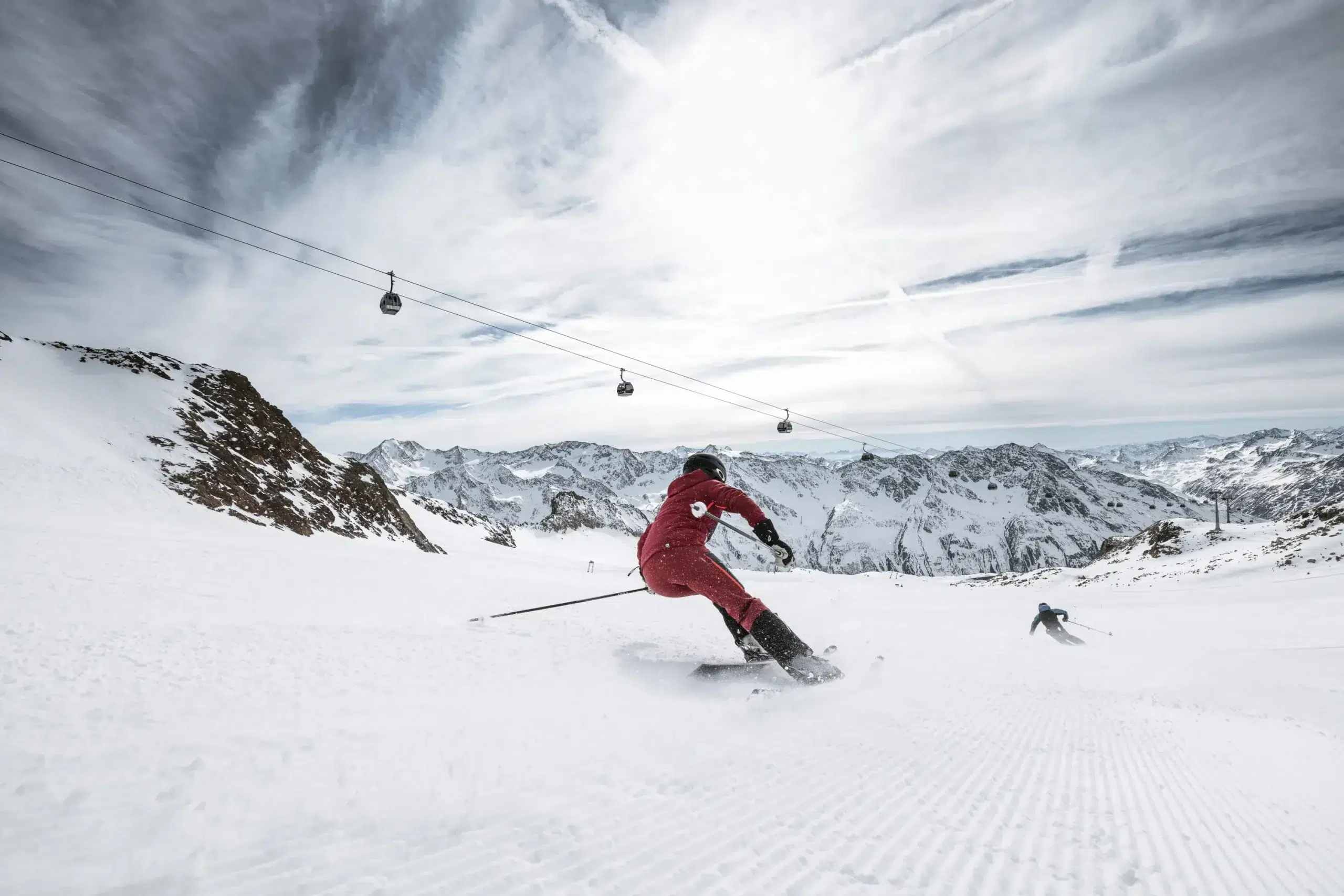 Zwei Skifahrer fahren während eines Skitests bei bewölktem Himmel einen verschneiten Hang hinunter, die Lifte sind über ihnen.