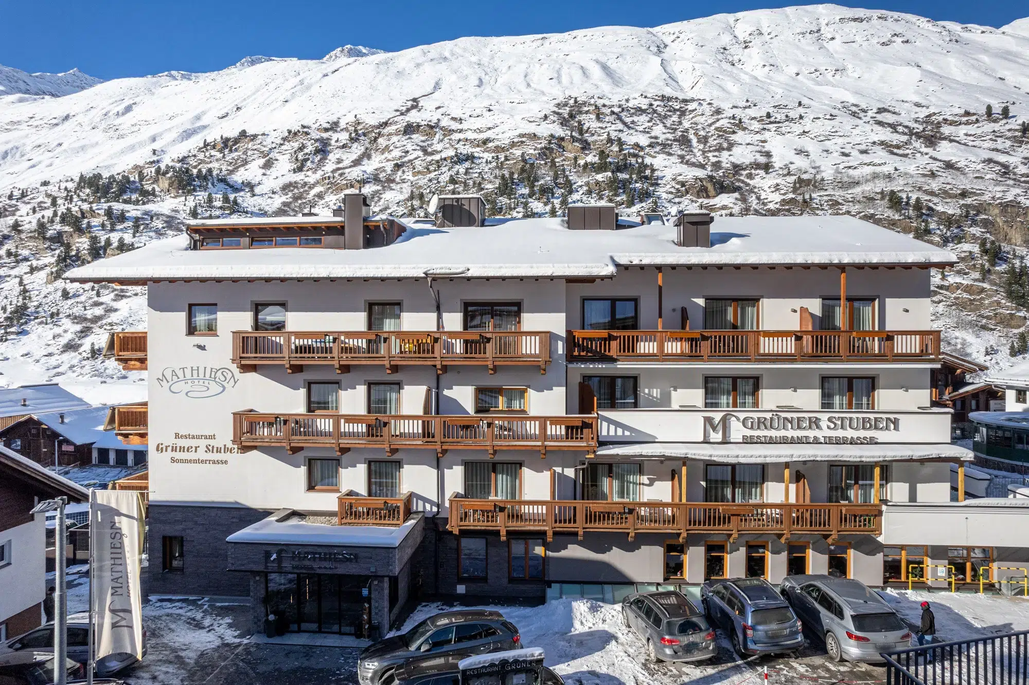 Verschneites Hotel Mathiesn mit Holzbalkonen, parkenden Autos und Bergen im Hintergrund.