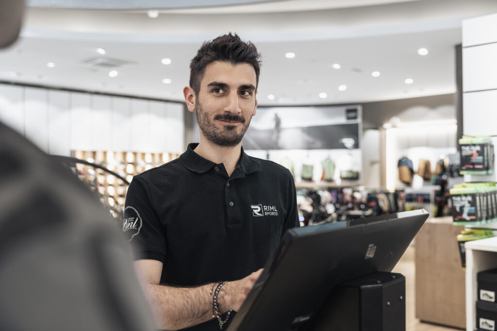 Ein Mann in einem schwarzen Poloshirt steht an einer Kasse und ruft Tipps zur Sicherheit am Berg.