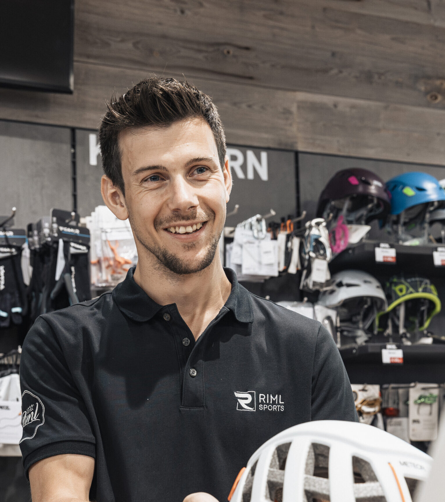Ein lächelnder Angestellter von Riml Sports Telfs überreicht einem Kunden in einem Sportgeschäft einen Fahrradhelm.