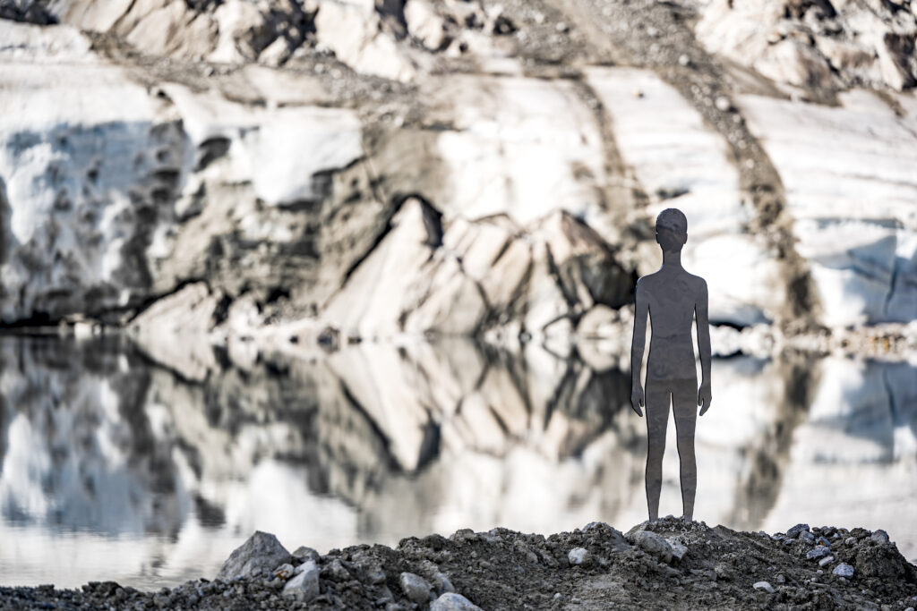 Silhouette eines Menschen, der vor einer felsigen, verschneiten Landschaft steht, die sich im Wasser spiegelt.