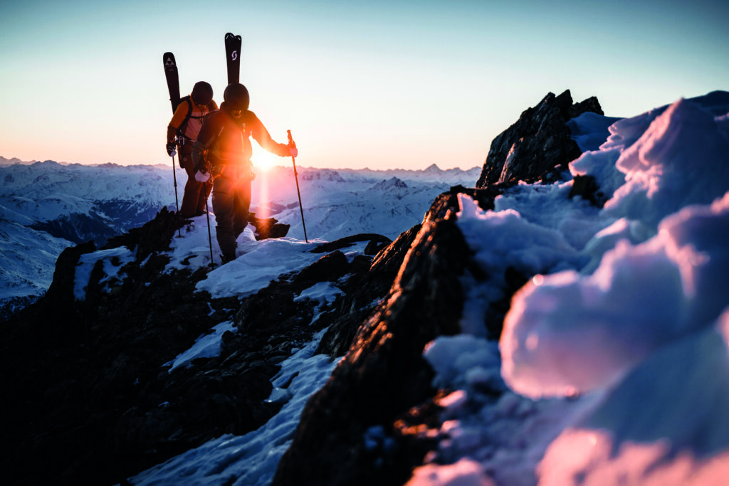 Zwei Menschen wandern bei Sonnenaufgang mit Skiern auf einen verschneiten Bergkamm.