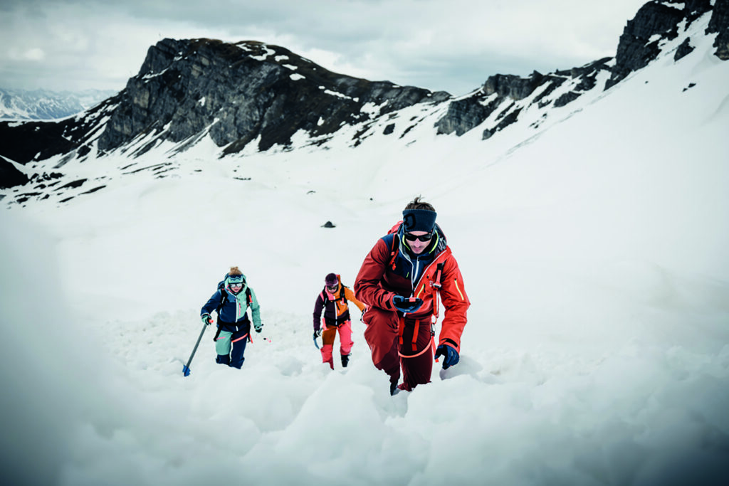 Drei Menschen in Winterkleidung wandern einen verschneiten Hang mit felsigen Gipfeln dahinter hinauf.