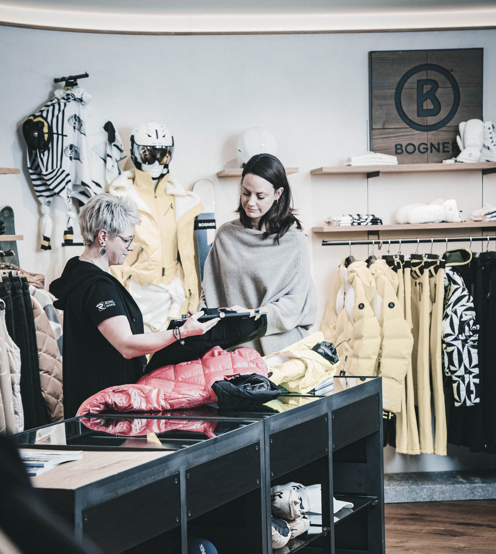 Zwei Frauen unterhalten sich am Schalter von Riml Sports Längenfeld, wo Jacken und Skiausrüstung ausgestellt sind.