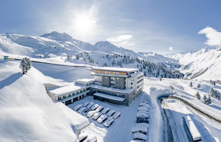 Hotel Rita nestled among snow-capped mountains, with cars parked under the bright winter sun.